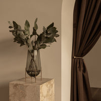 Elegantly displayed grey glass vase with dark green foliage, set against a warm beige wall and rich brown curtain.