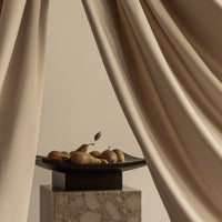 Beige curtains frame a dark plate with pears, set on a light marble pedestal, creating a soft, elegant aesthetic.