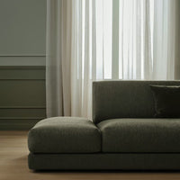 Gray upholstered sofa with a minimalist design, set against light sheer curtains and a muted green wall.