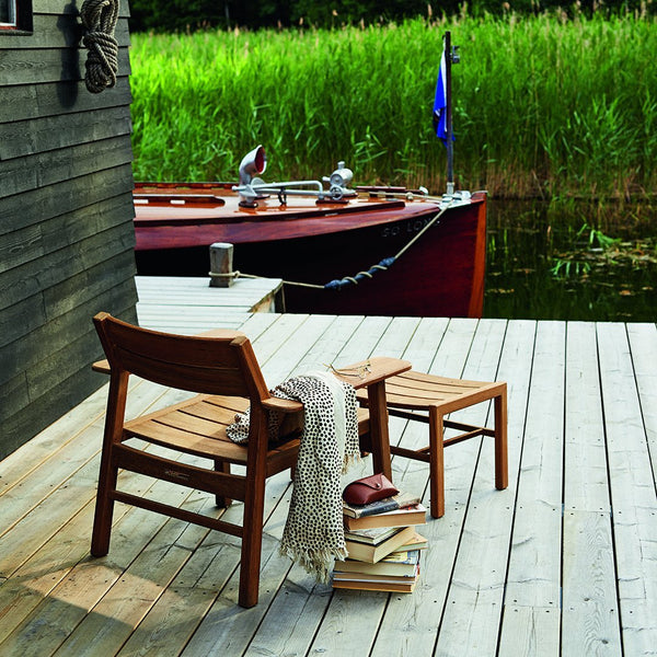 "Natural wood chairs on a light deck with green grass, blue water, and a red boat backdrop."