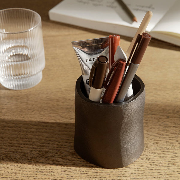 Dark brown ceramic pen holder with glossy finish, featuring a textured design, surrounded by pens and a clear glass.