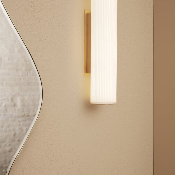 Modern wall sconce in soft white and gold, featuring a sleek ribbed design against a neutral beige wall.