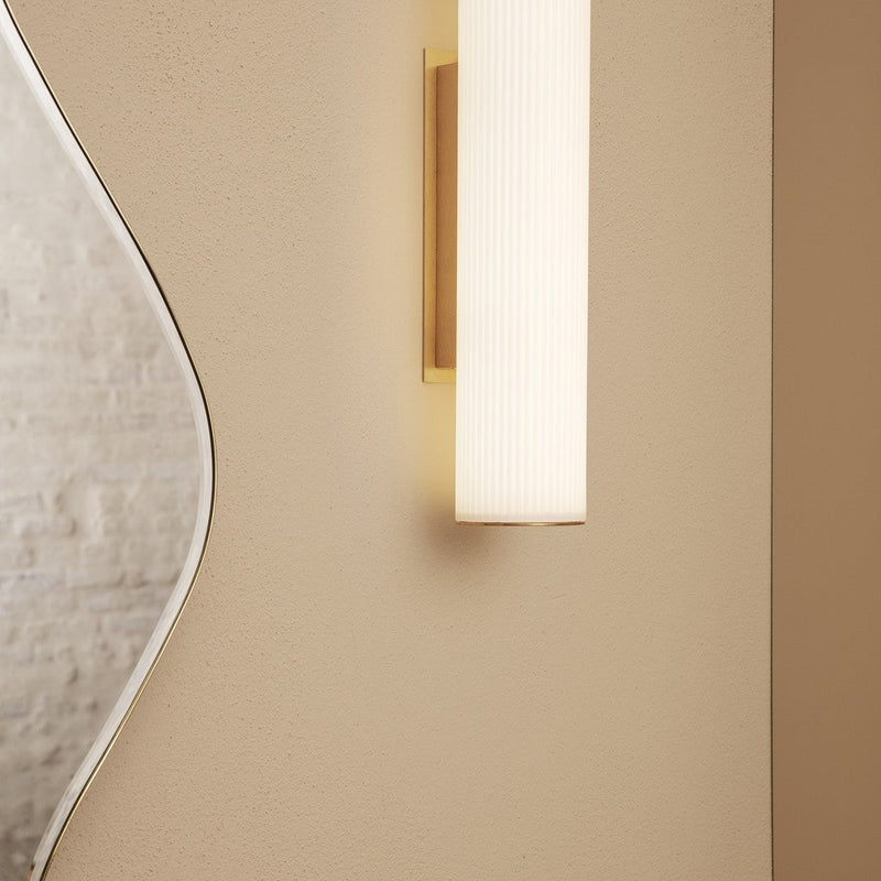 Modern wall sconce in soft white and gold, featuring a sleek ribbed design against a neutral beige wall.