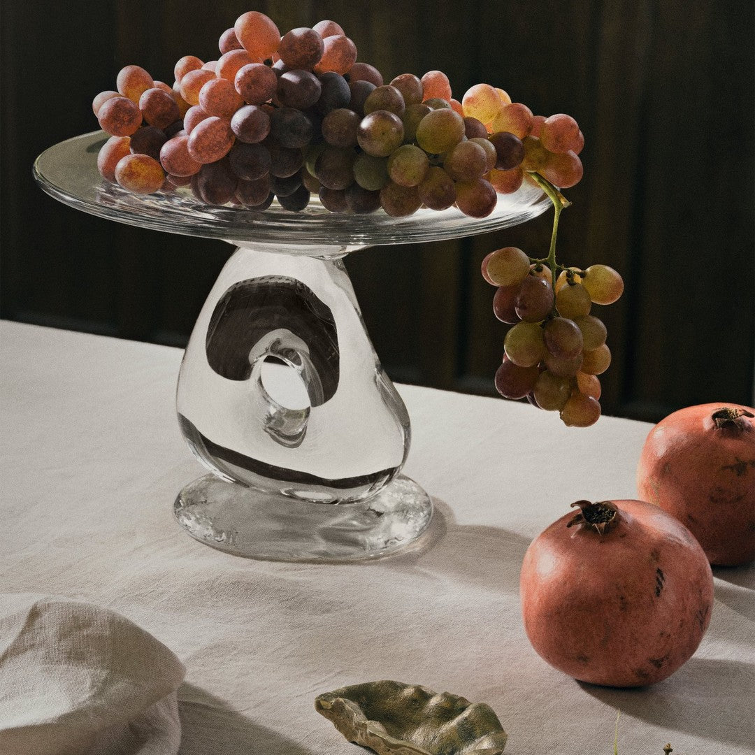 "Stylish glass cake stand with a unique shape, showcasing clusters of purple and green grapes with red pomegranates."