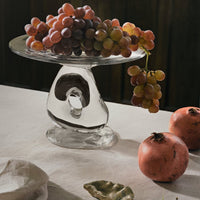 "Stylish glass cake stand with a unique shape, showcasing clusters of purple and green grapes with red pomegranates."