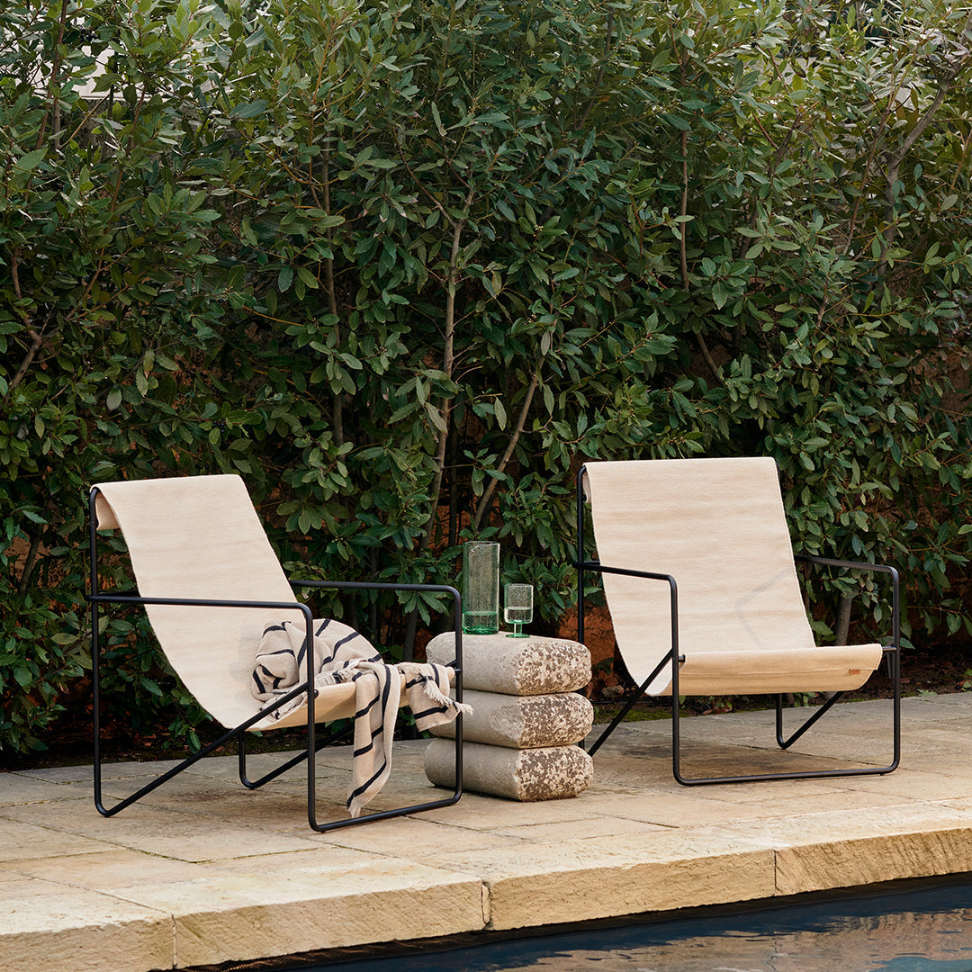 Two light beige chairs with black frames and patterned throw, placed next to stacked pillows near a pool amidst greenery.