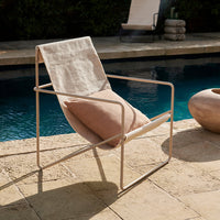 Beige fabric lounge chair with a light metal frame, featuring a textured cushion, set against a blue pool backdrop.