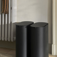 Black cylindrical bins with a sleek matte finish, placed beside a light-colored wall and a striped fabric.