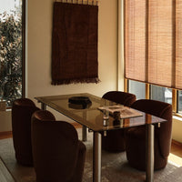 Modern dining room featuring brown upholstered chairs, a glass table, and a textured wall hanging with earthy tones.