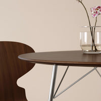 Brown wood table with a sleek round top, paired with a sculptural chair silhouette against a soft beige backdrop.