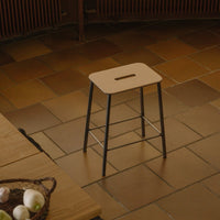 Light tan wooden stool with a rectangular cutout, paired with black metal legs against a rustic tile floor.