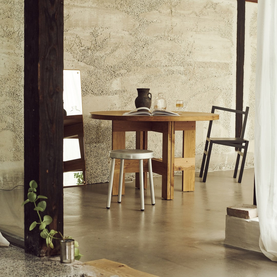 "Modern wooden dining table with a natural finish, paired with a silver metal stool and black chair against a textured wall."