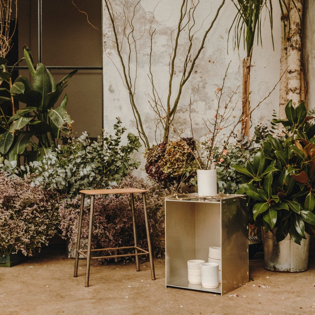 "Metallic silver storage cube with white candle, surrounded by lush greenery and soft pink floral accents."