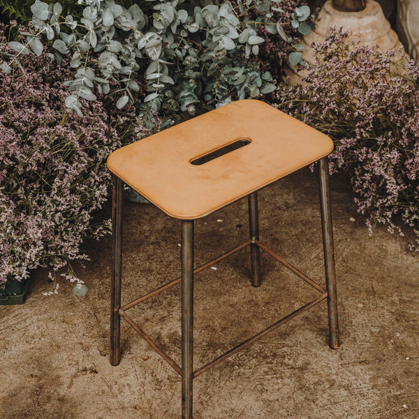 Tan wooden stool with a sleek design and metallic legs, surrounded by greenery and flowers.