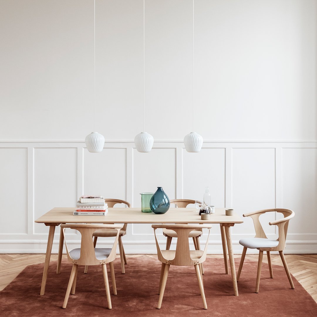 Light wood dining table with beige and white chairs on a rich brown rug, complemented by soft white pendant lights.