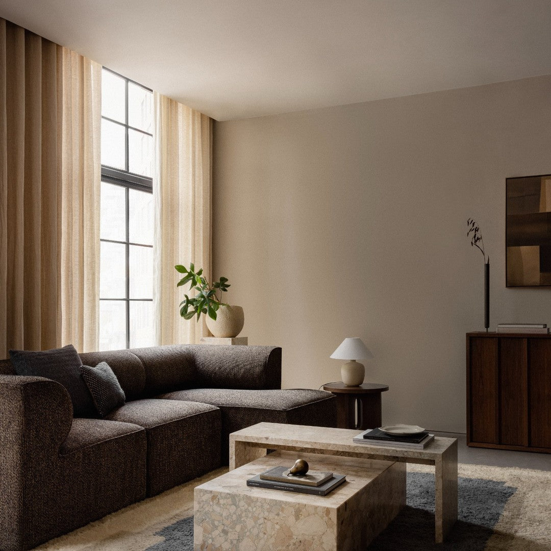 Earthy brown sofa with subtle textures, cream curtains, light beige walls, and a natural stone coffee table.