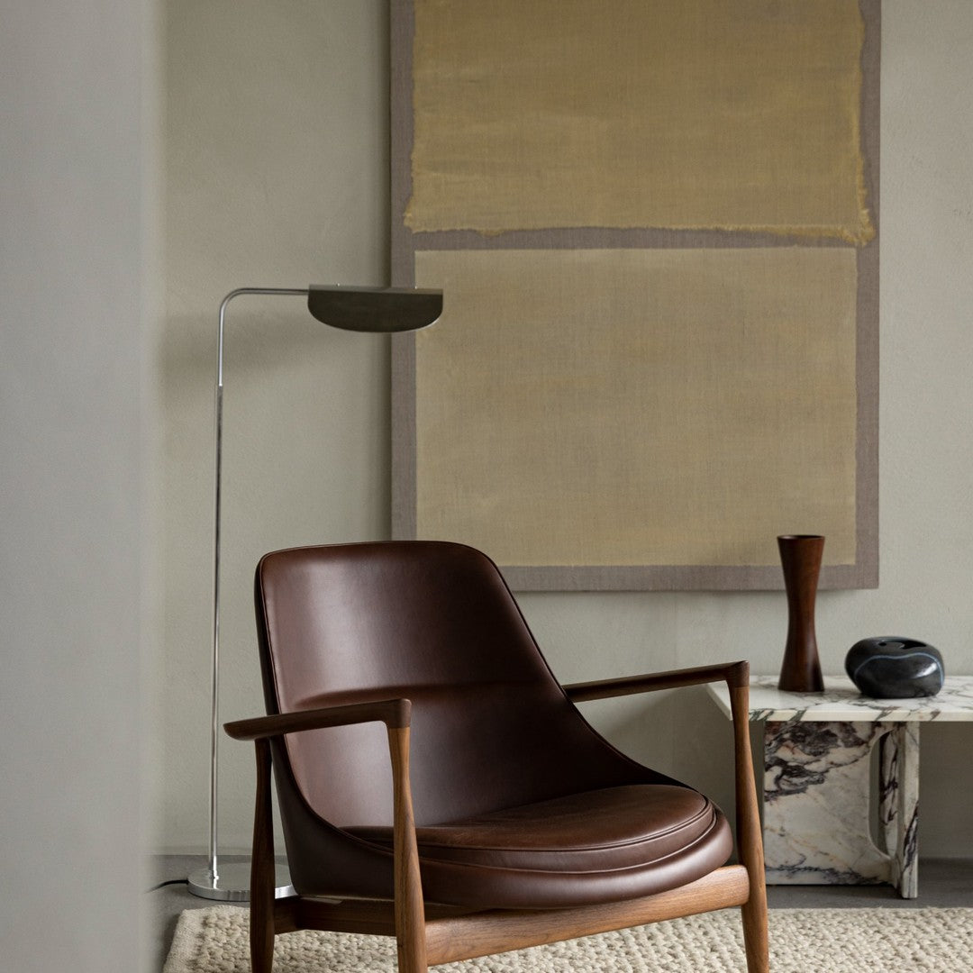 Brown leather armchair with sleek wooden frame, set against a neutral backdrop and textured beige wall art.