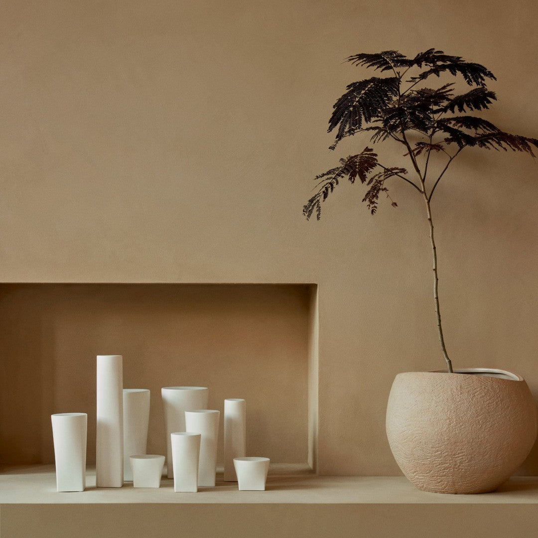 White ceramic vases in various shapes and sizes, set against a warm, neutral backdrop with a textured planter.