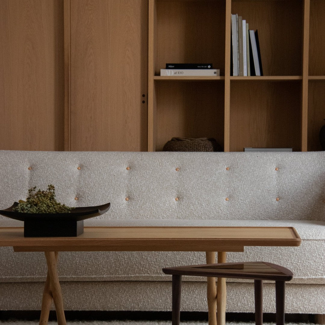 Beige textured sofa with button detailing, paired with a light wooden table and dark wood stool, set against a natural backdrop.