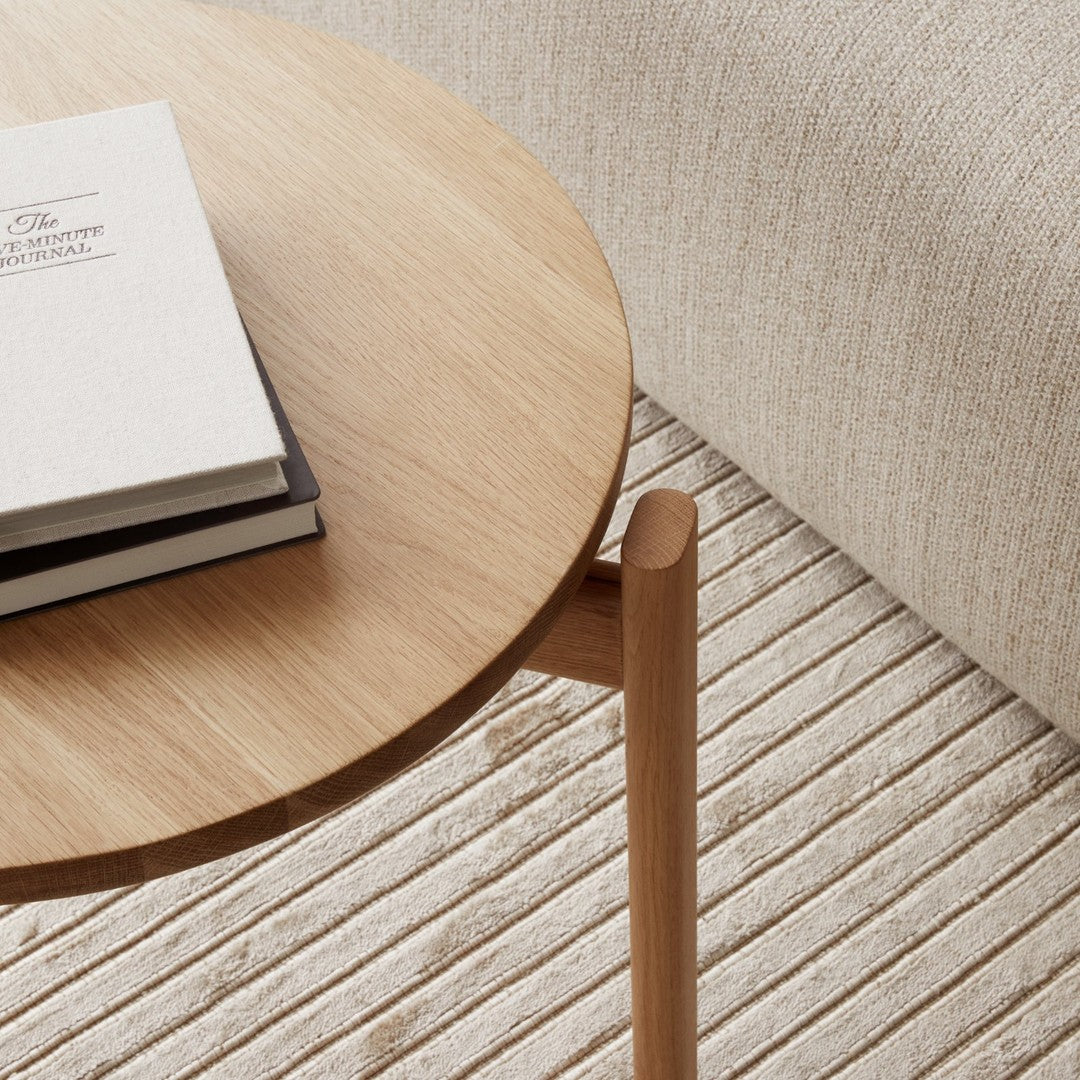 Light oak round table top, natural wood grain, against textured beige fabric background and striped cream rug.