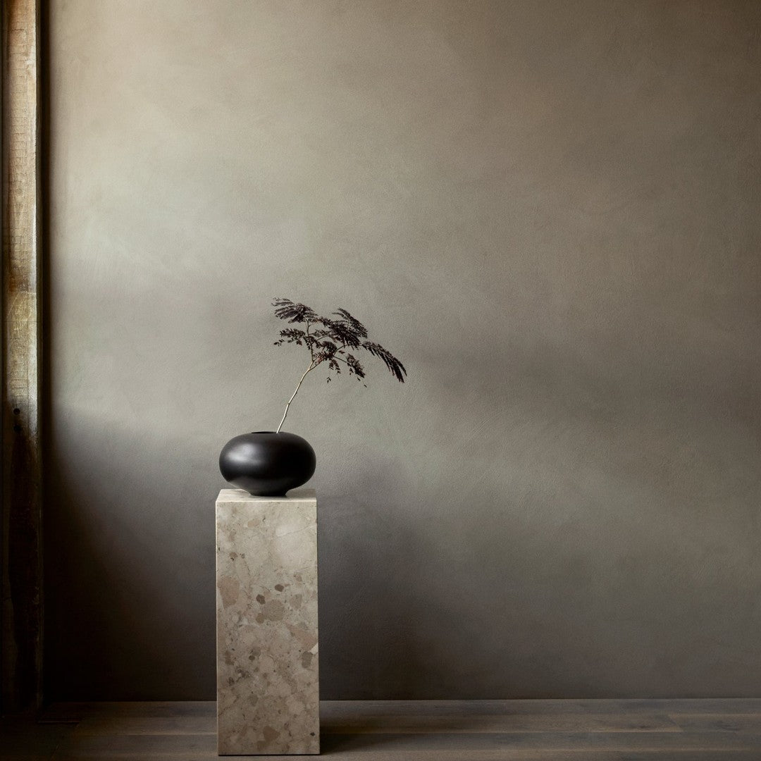 Black stone vase atop a textured beige pedestal, with minimalistic brown wall background and delicate brown plant stems.