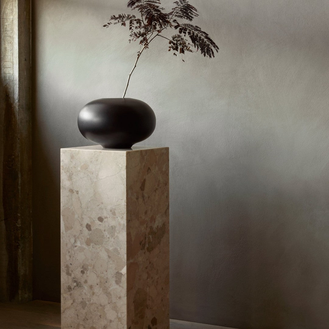 Black ceramic vase on a textured beige pedestal against a soft gray background. Elegant minimalist design.