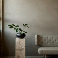 Elegant green leaves in a black vase atop a beige marble pedestal, complemented by a light gray tufted sofa against a muted backdrop.