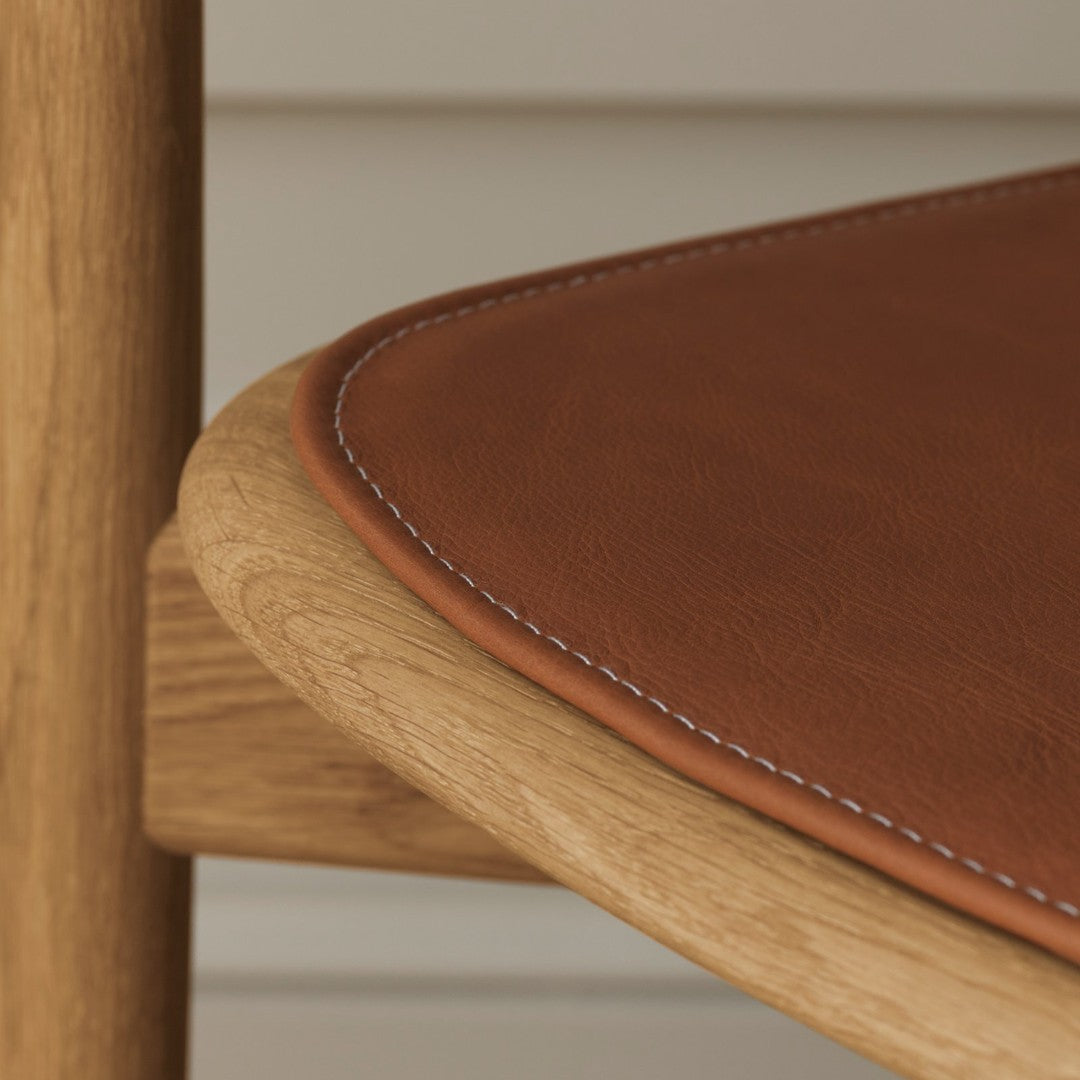 Brown leather cushion with white stitching on a natural wood chair, featuring a sleek, minimalist design.