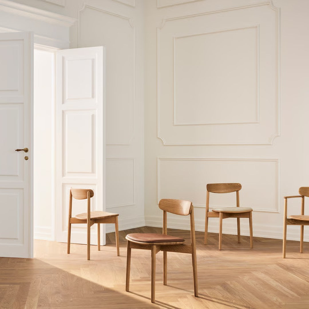 Four minimalist wooden chairs in natural finish, set against white walls and a light wood floor.