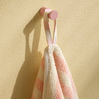 Soft beige towel with coral pink stripes, hanging from a pastel pink hook against a yellow wall.