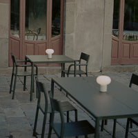 Minimalist outdoor dining setup with black metal tables and chairs, featuring a round white lamp. Rustic stone background.
