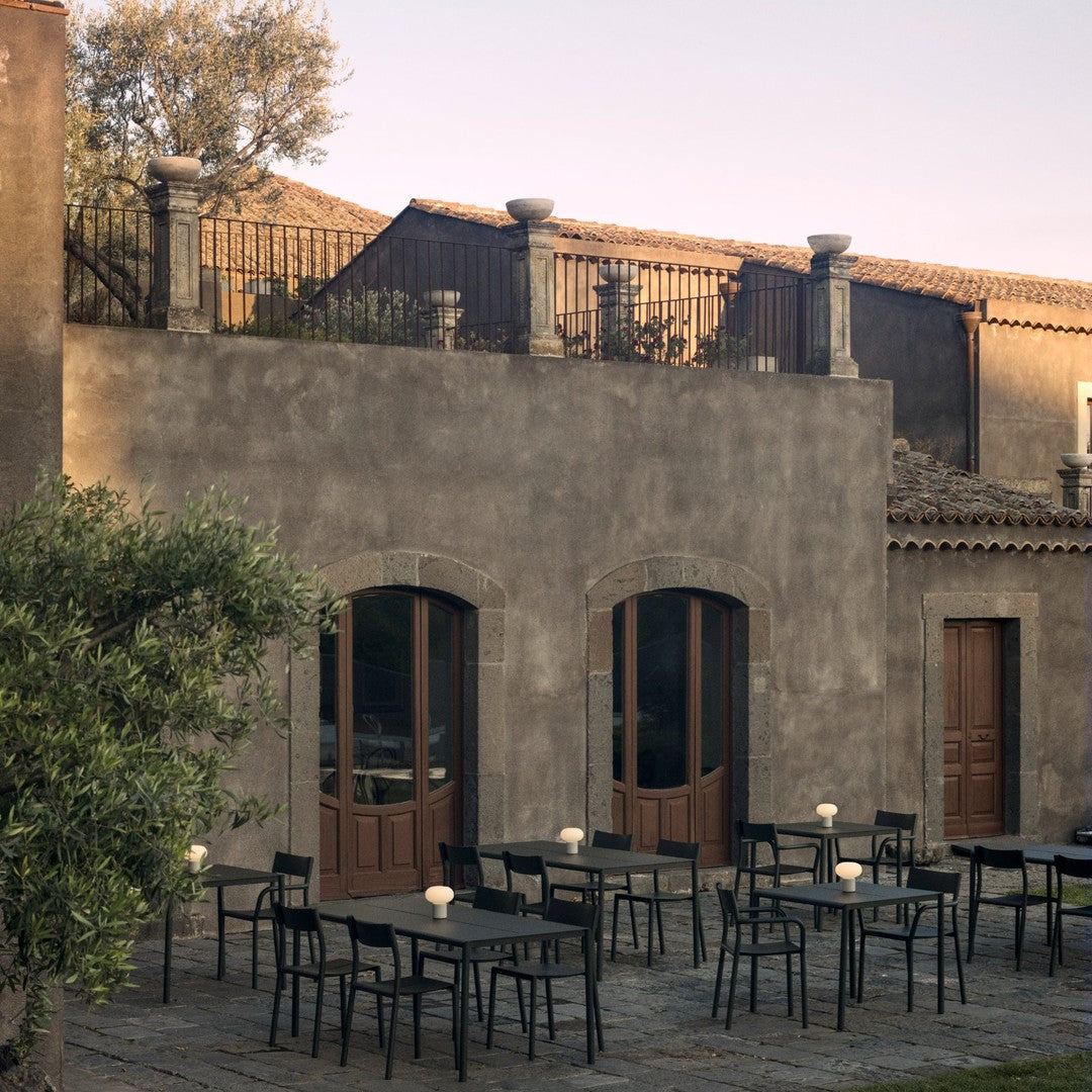 "Outdoor seating with black tables, minimalist design against a rustic gray stone wall and soft greenery."