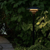Black outdoor lamp with a golden shade, surrounded by lush green foliage and stone pathway. Soft, warm glow at dusk.
