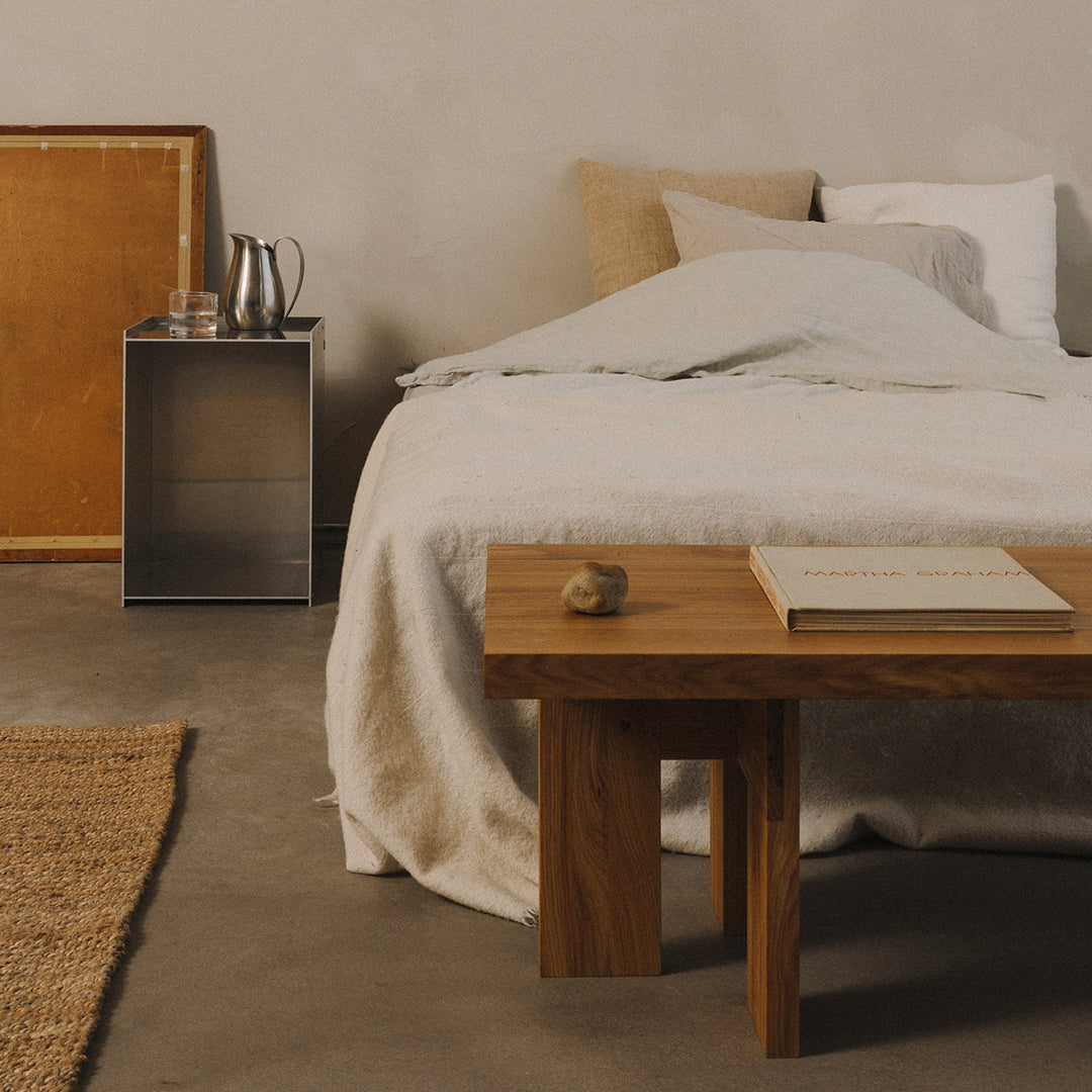 Neutral beige and soft gray bedding with a textured white throw, complemented by warm wood tones and a woven rug.