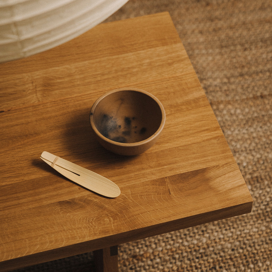 Brown ceramic bowl with dark speckles beside a light wooden spoon on a natural wood table, with a textured woven background.