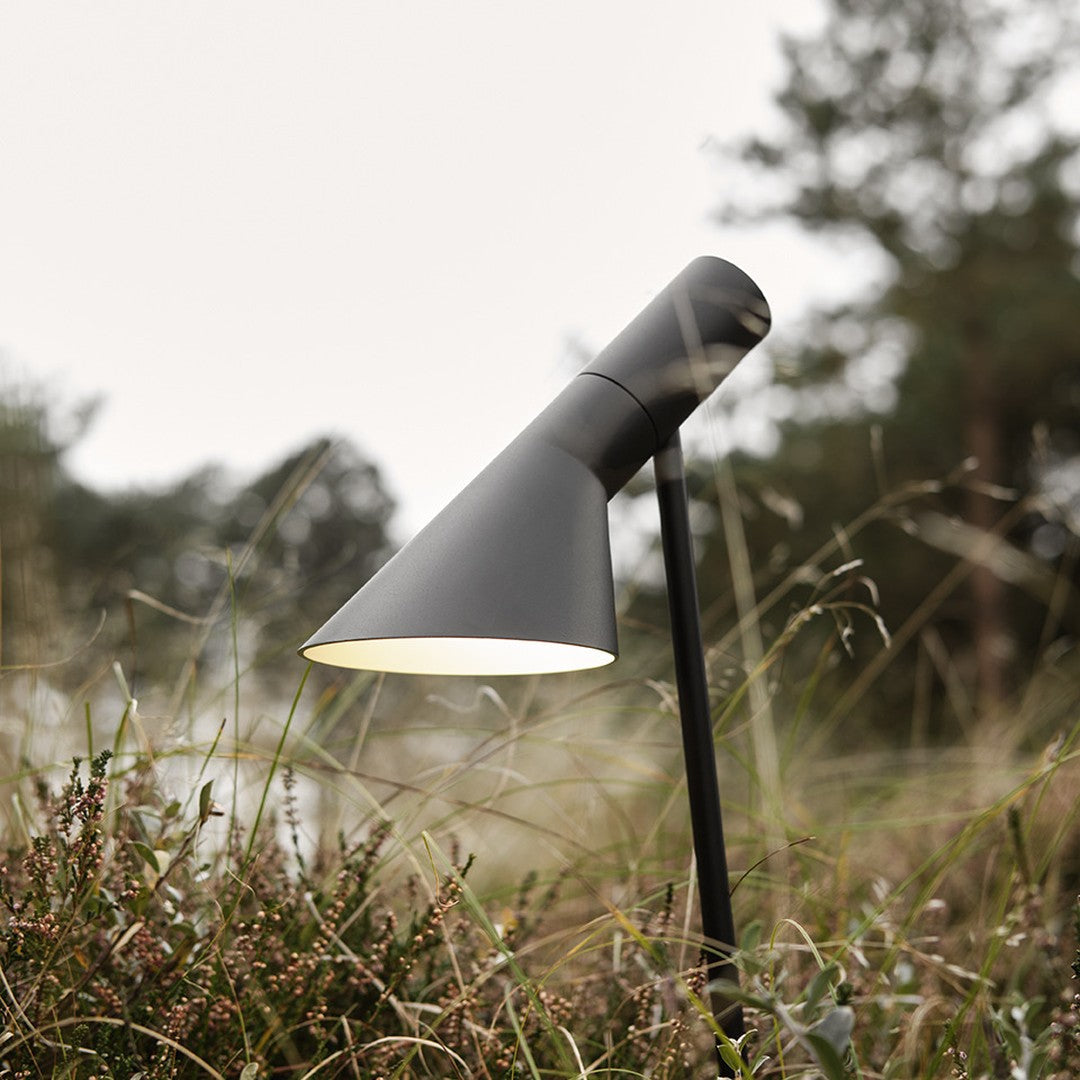 Black outdoor lamp with a sleek conical shade, set against a natural backdrop of green grass and soft blurred trees.