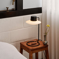 Modern black and white lamp with a circular shade on a wooden stool, accented by a minimalist glass vase and yellow flower.