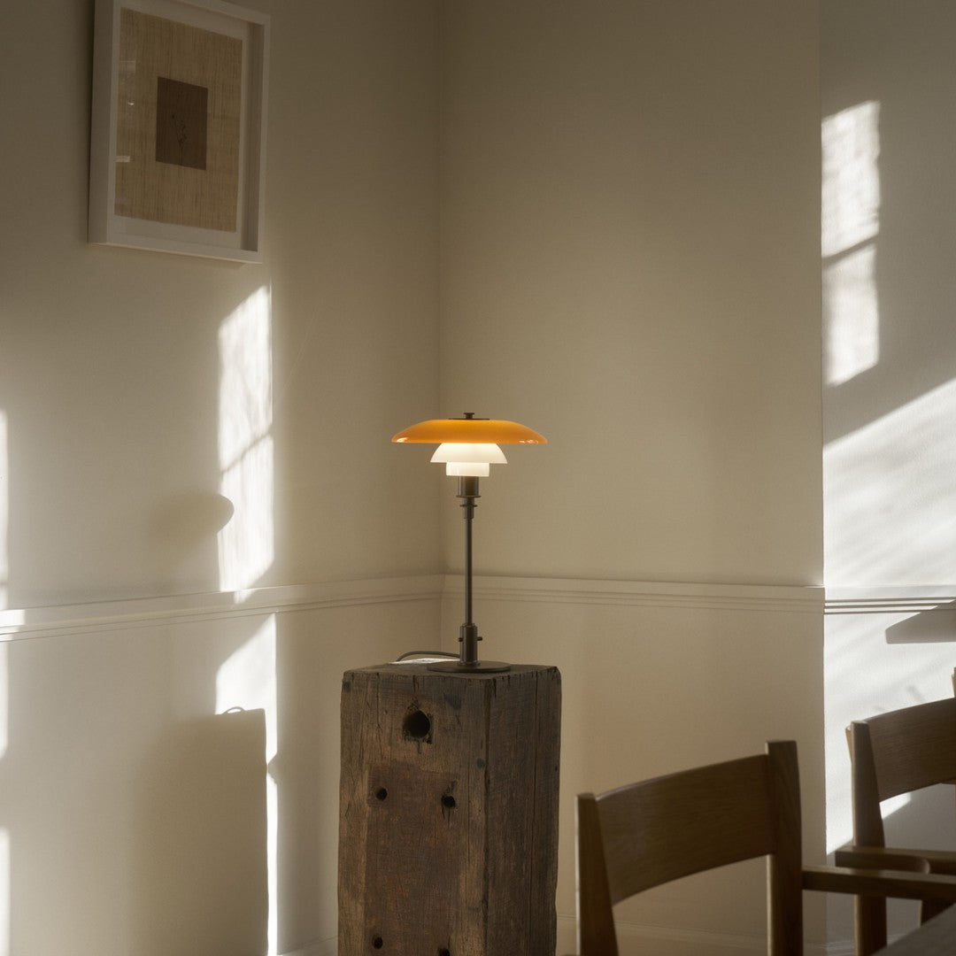 Orange and white lamp with a round shade, placed on a wooden block in a sunlit, minimalist room.