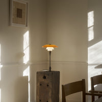Orange and white lamp with a round shade, placed on a wooden block in a sunlit, minimalist room.