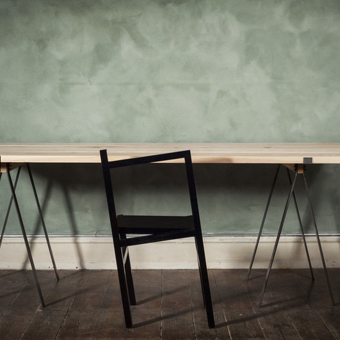 Minimalist black chair against a textured green wall with a light wooden table, showcasing modern design elements.