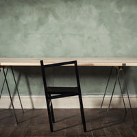 Minimalist black chair against a textured green wall with a light wooden table, showcasing modern design elements.