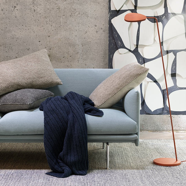 Light blue sofa with gray and navy pillows, navy throw blanket, and orange lamp against a textured, abstract wall.