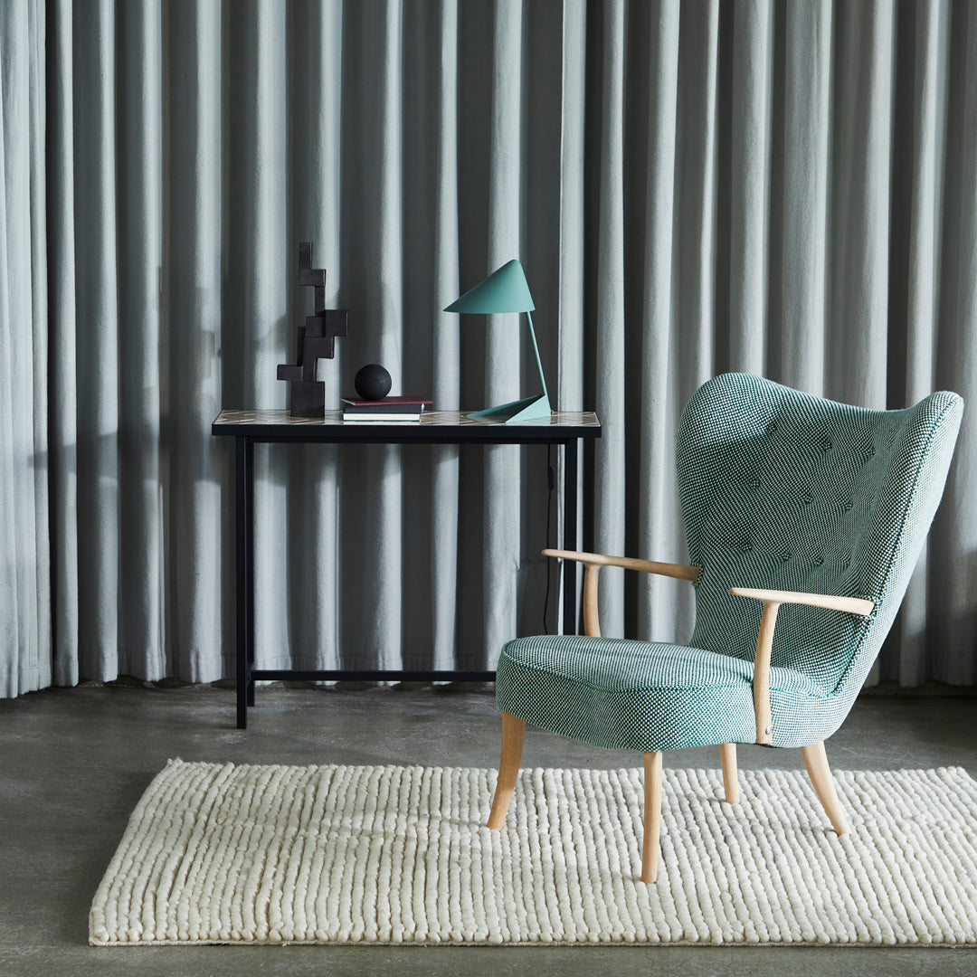 "Soft teal upholstered chair with wooden legs, set against gray curtains and a textured white rug."