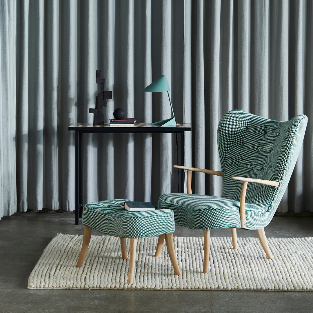 Teal upholstered armchair with matching ottoman, featuring subtle tufted texture, against gray curtains and a neutral rug.