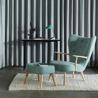 Teal upholstered armchair with matching ottoman, featuring subtle tufted texture, against gray curtains and a neutral rug.
