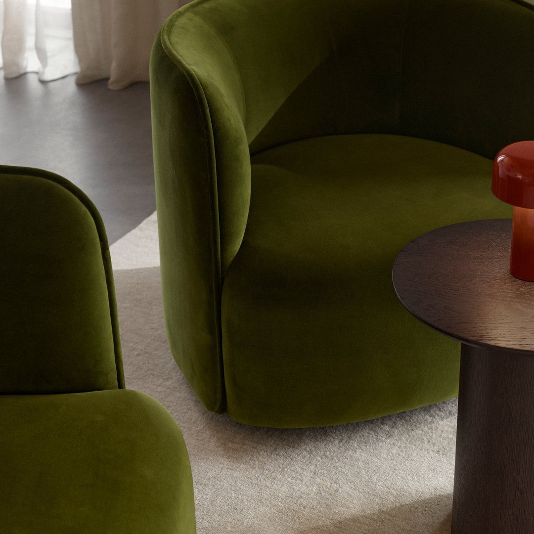 Velvet green armchairs with a smooth finish, paired with a round dark wood table and a vibrant orange accent piece.